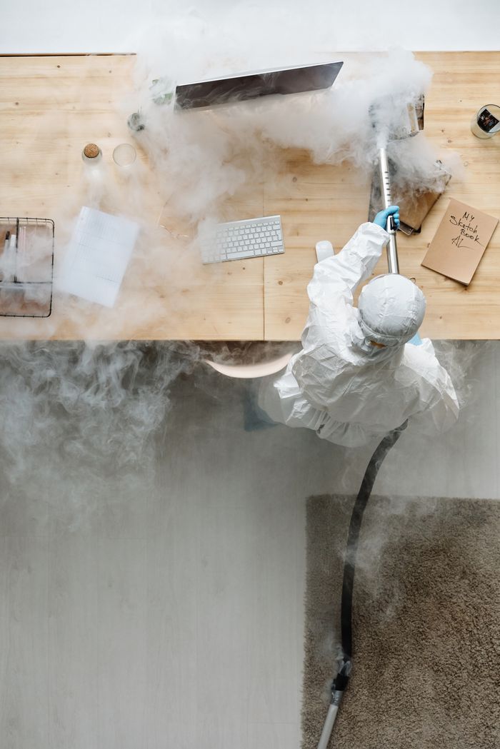 services-03 A person in protective gear sanitizing an office desk using fumigation equipment during daytime.
