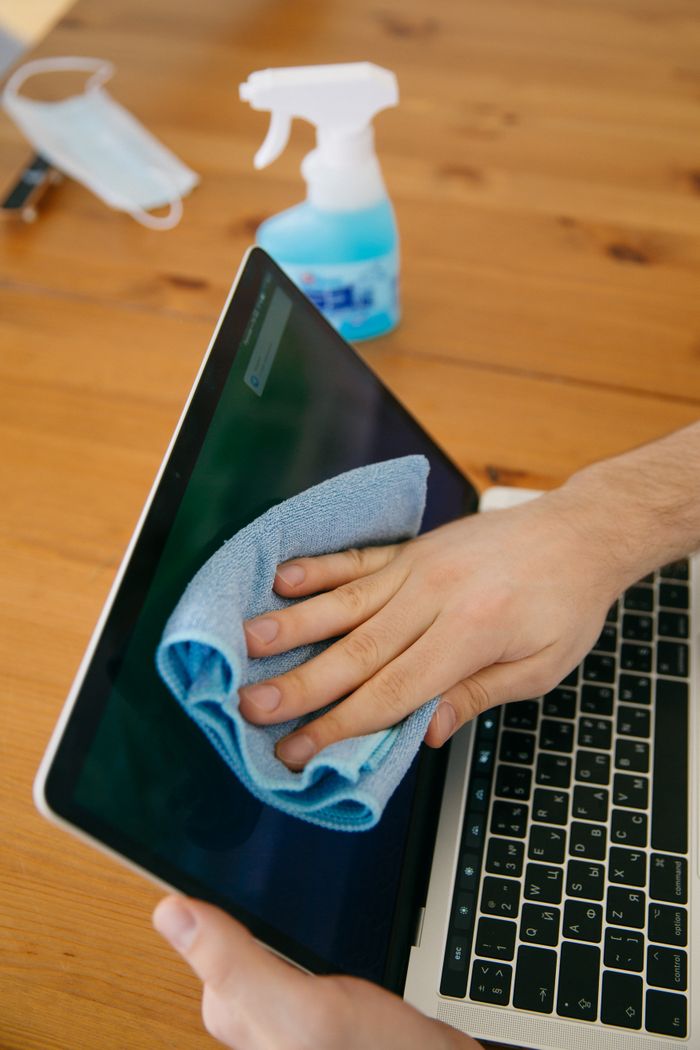why-choose-us A person wipes a laptop screen using a blue cloth and spray bottle on a wooden table for hygiene.