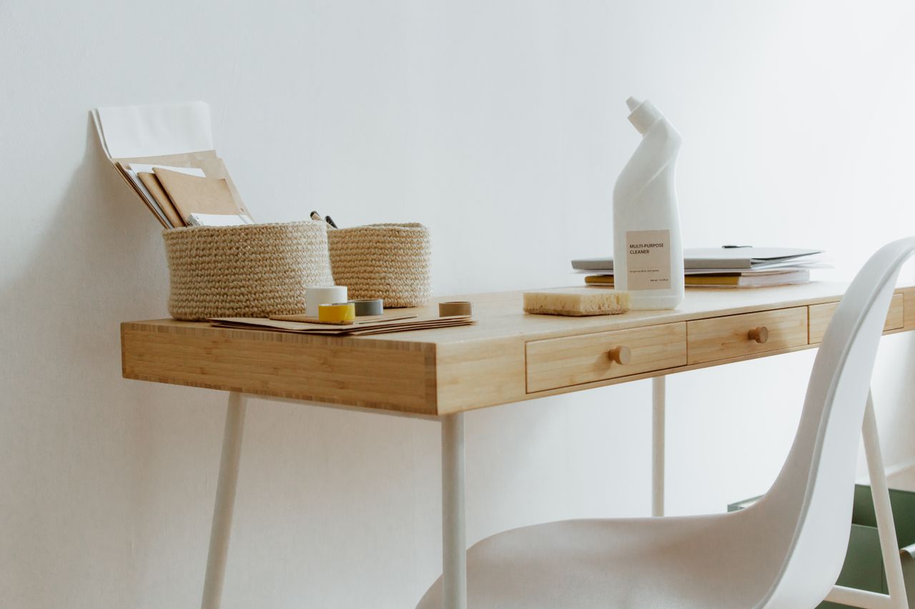 gallery-1 A clean and organized home office desk with cleaning products, papers, and storage baskets.